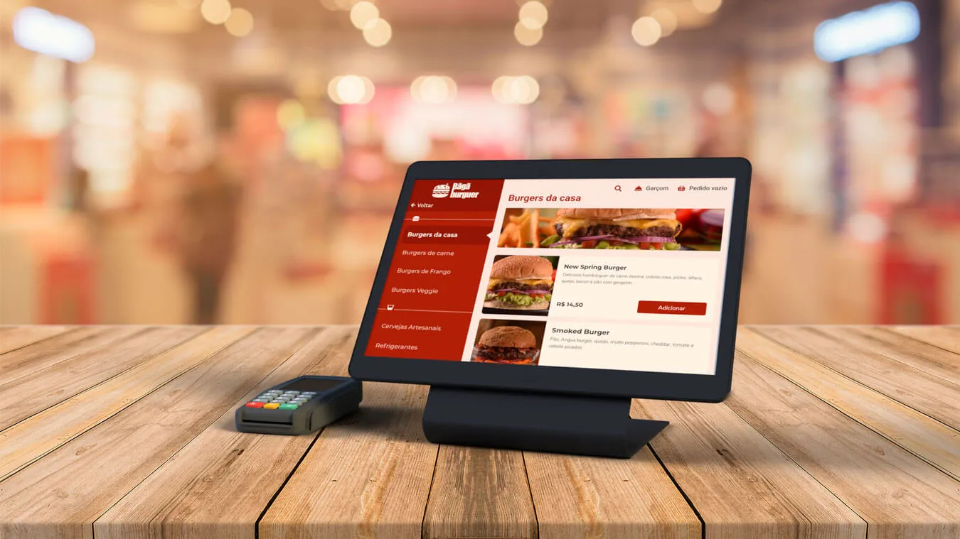 Tablet e maquininha de cartão de crédito em cima de uma bancada de madeira com o fundo de um restaurante desfocado